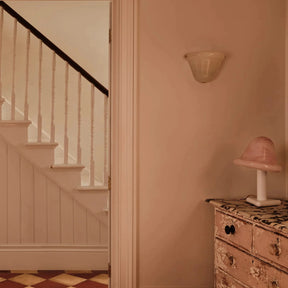 A warm, vintage-style interior scene featuring a dusty pink wall, a white staircase with a dark wooden railing on the left, and a distressed pink and gold chest of drawers with a matching pink lamp on the right.