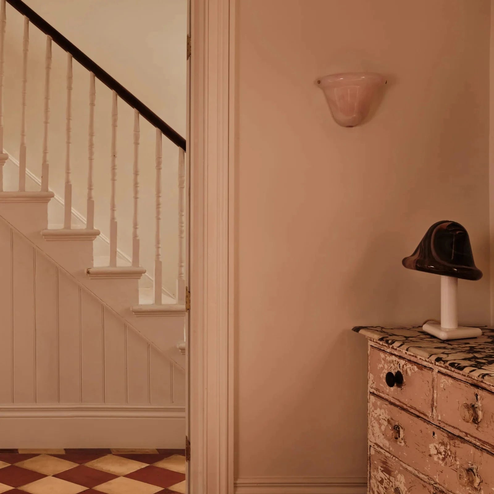 A warm-toned interior hallway scene with a cream-colored wall sconce, a white staircase with a dark handrail on the left, and a distressed pinkish-beige dresser with a small black hat on top in the foreground.