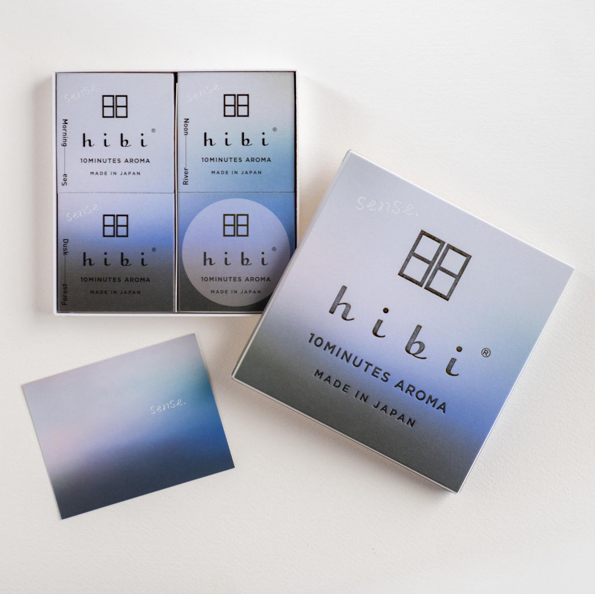 A clean studio flat-lay of a hibi Tominutes Aroma product set on a white background, featuring a main outer box, a smaller inner box with two sachets, a round sachet, and a small card with a blue-to-gray gradient.
