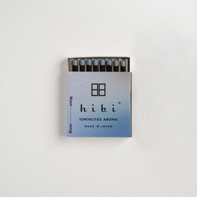 A matchbox of Hibi 10minutes Aroma matches mounted on a white textured wall. The matchbox features a light blue and white gradient cover with the Hibi brand logo and name, and a small portion of the dark wooden matches is visible at the top.