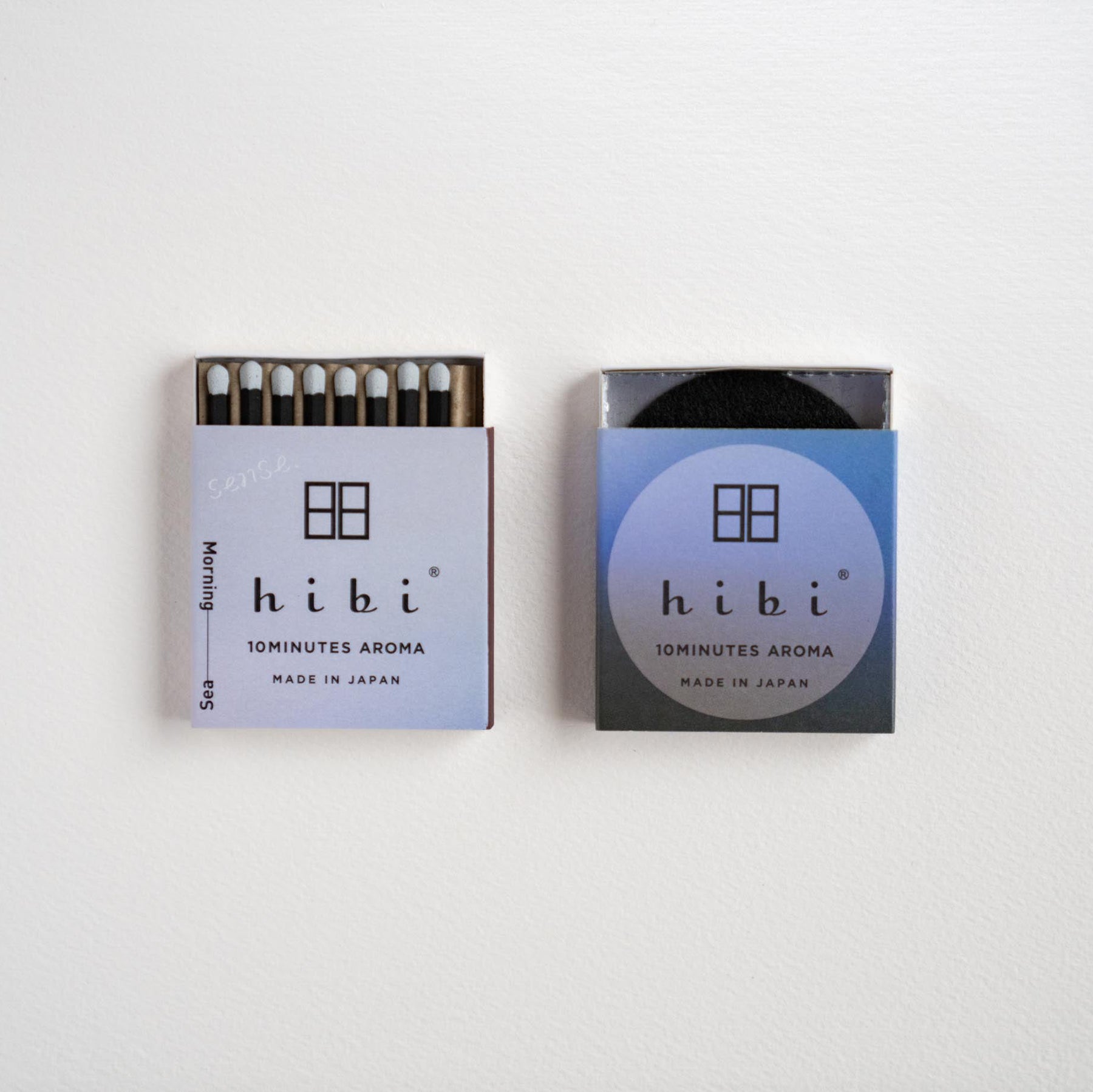 Two Hibi matchboxes side by side on a plain white background. The left matchbox is open, showing the dark match heads and matches inside, while the right matchbox is closed with a blue gradient label.