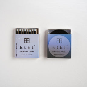Two Hibi matchboxes side by side on a plain white background. The left matchbox is open, showing the dark match heads and matches inside, while the right matchbox is closed with a blue gradient label.