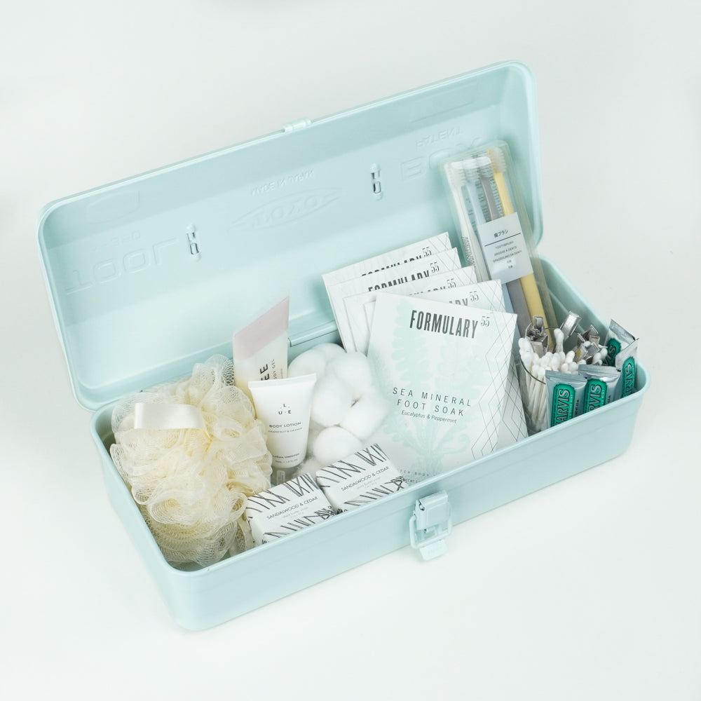 Open light blue Toyo Steel Y-350 metal toolbox styled as a self-care kit, holding bath and grooming items such as loofah, cotton balls, soap, foot soak packets, cotton swabs, and bamboo toothbrushes on a white background.