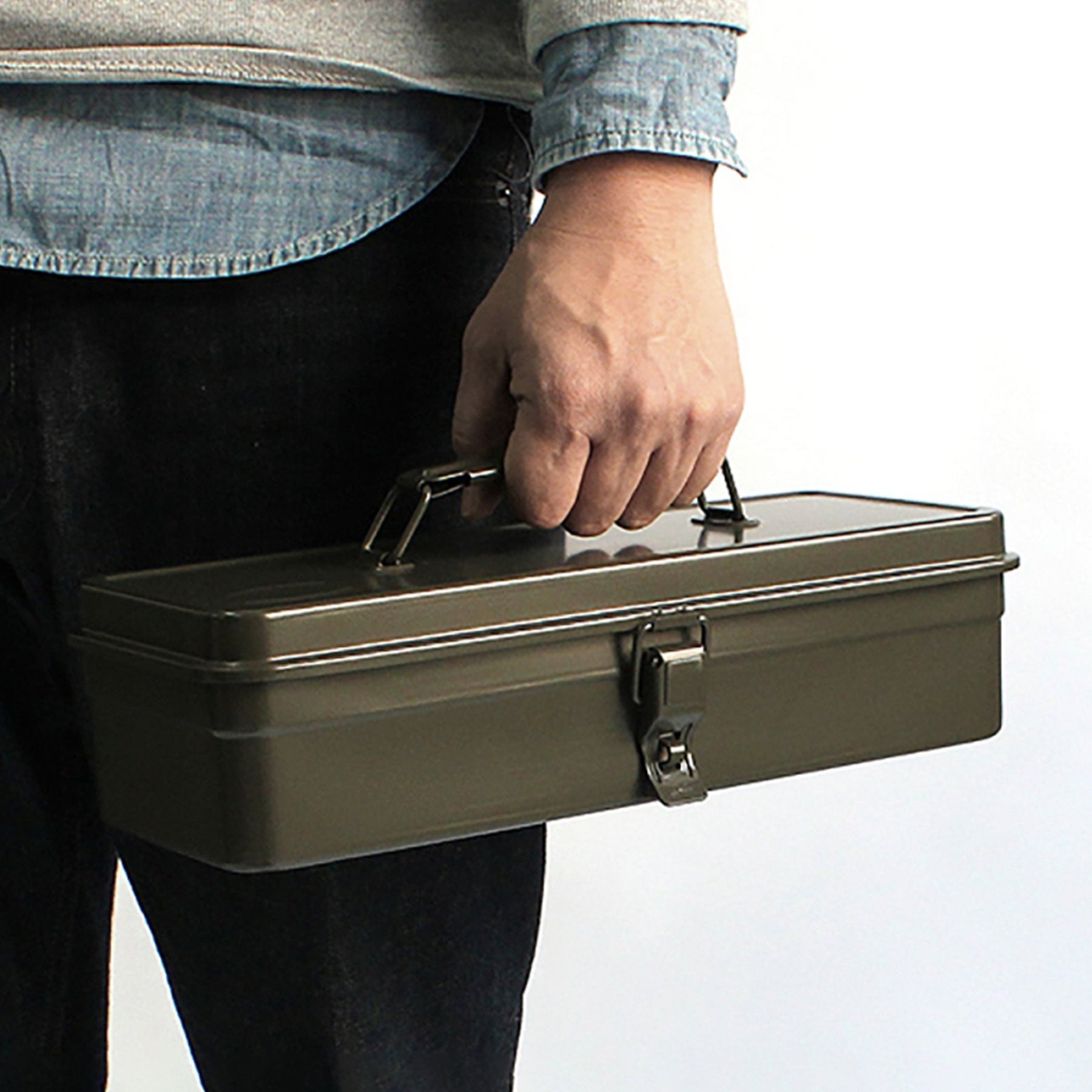 Person holding a military green Toyo Steel T-320 metal toolbox by its handle, shown at waist level against a white background.