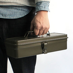 Person holding a military green Toyo Steel T-320 metal toolbox by its handle, shown at waist level against a white background.
