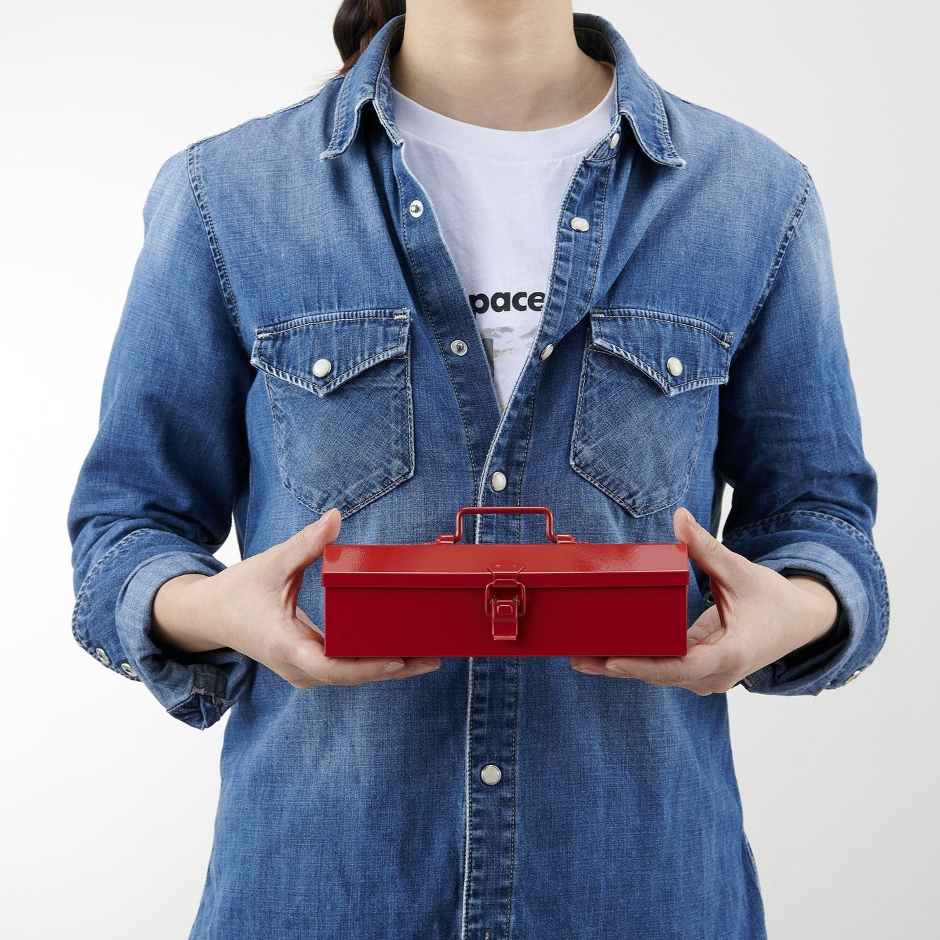 Front view of a person wearing a denim shirt and white T-shirt, holding a small red Toyo Steel Y-17 metal toolbox with a top handle and front latch, shown at chest height against a clean white background.